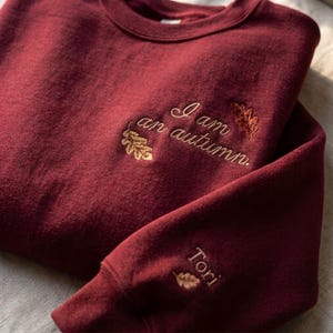 May include: A deep burgundy crewneck sweatshirt featuring gold embroidered oak leaves and the text "I am an autumn." on the chest. The sleeve cuff displays the name "Tori" embroidered in gold thread above a small oak leaf design.