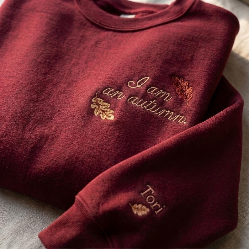 May include: A deep burgundy crewneck sweatshirt featuring gold embroidered oak leaves and the text "I am an autumn." on the chest. The sleeve cuff displays the name "Tori" embroidered in gold thread above a small oak leaf design.