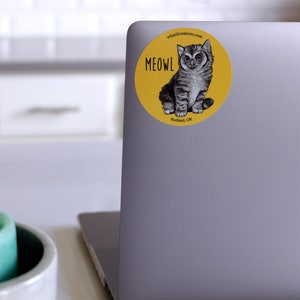 Pode incluir: Um adesivo circular amarelo com uma ilustra&ccedil;&atilde;o em preto e branco de um gato com olhos grandes. O texto "MEOWL" est&aacute; escrito em letras pretas no adesivo. Os textos "whatifcreations.com" e "Portland, OR" tamb&eacute;m est&atilde;o escritos no adesivo.