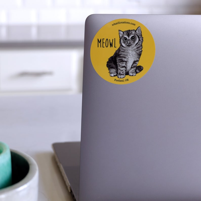 Pode incluir: Um adesivo circular amarelo com uma ilustra&ccedil;&atilde;o em preto e branco de um gato com olhos grandes. O texto "MEOWL" est&aacute; escrito em letras pretas no adesivo. Os textos "whatifcreations.com" e "Portland, OR" tamb&eacute;m est&atilde;o escritos no adesivo.
