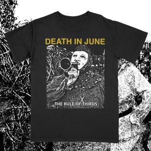 Może przedstawiać: Czarny t-shirt z czarno-białą grafiką przedstawiającą osobę w masce trzymającą megafon. Złoty napis "DEATH IN JUNE" znajduje się nad obrazem, a pod nim "THE RULE OF THIRDS". Ciemny, artystyczny design.