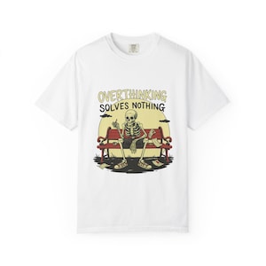 Puede incluir: Camiseta blanca con un diseño de esqueleto de dibujos animados sentado en un banco rojo, fumando un cigarrillo. El texto "OVERTHINKING SOLVES NOTHING" está encima. Papeles esparcidos alrededor del banco, sobre un fondo amarillo.