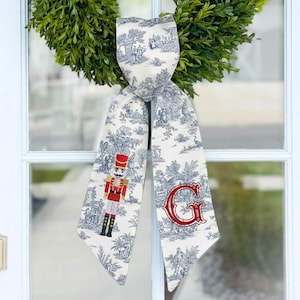 May include: A decorative bow with a blue and white toile pattern, featuring a red embroidered letter "G" and a nutcracker figure. The bow is attached to a green wreath, creating a festive holiday decoration.