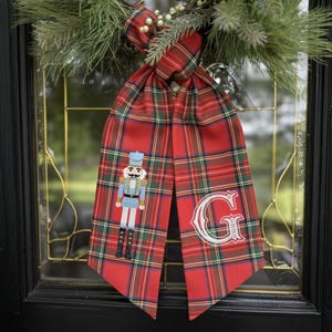 May include: A festive Christmas door decoration featuring a red plaid ribbon with a nutcracker embroidery and a white embroidered letter "G". The ribbon is adorned with a pine and berry wreath.