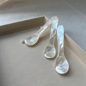 May include: Three iridescent mother-of-pearl spoons with a pearlescent sheen. The spoons are arranged in a group, with one spoon partially visible. A small pearl rests near the spoons. The spoons are set in a light-colored box.