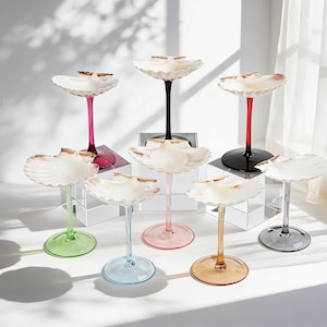 May include: A collection of decorative glass stands with seashell-shaped tops. Each stand features a different colored stem and base, including pink, black, red, green, blue, peach, and gray. The stands are arranged on a white surface.