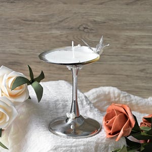 Puede incluir: Un portavelas plateado con un adorno de mariposa, que sostiene una vela blanca. El portavelas está sobre una tela blanca, rodeado de rosas blancas y melocotón. El fondo es una superficie de madera.