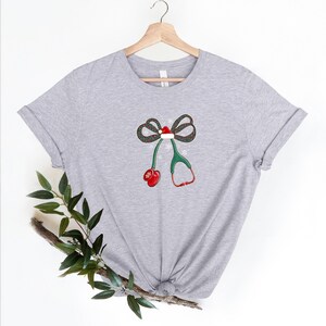 Puede incluir: Camiseta gris claro con un dise&ntilde;o de estetoscopio con tem&aacute;tica navide&ntilde;a. El dise&ntilde;o incluye un lazo con un gorro de Pap&aacute; Noel, tubos de estetoscopio verdes y auriculares rojos. La camiseta est&aacute; colgada en una percha de madera.