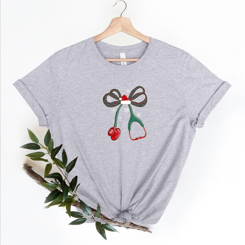Puede incluir: Camiseta gris claro con un dise&ntilde;o de estetoscopio con tem&aacute;tica navide&ntilde;a. El dise&ntilde;o incluye un lazo con un gorro de Pap&aacute; Noel, tubos de estetoscopio verdes y auriculares rojos. La camiseta est&aacute; colgada en una percha de madera.