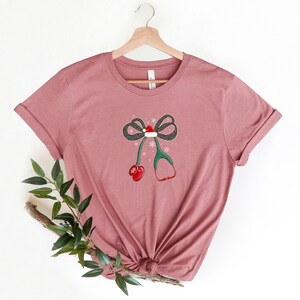 Puede incluir: Una camiseta rosa polvoriento con un dise&ntilde;o festivo. El gr&aacute;fico incluye un lazo con un gorro de Pap&aacute; Noel, copos de nieve y un estetoscopio con detalles rojos. Ideal para profesionales de la salud durante la temporada navide&ntilde;a.