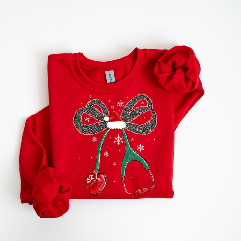 Puede incluir: Sudadera roja con un dise&ntilde;o de estetoscopio con tem&aacute;tica navide&ntilde;a. El dise&ntilde;o presenta un lazo con un gorro de Pap&aacute; Noel, copos de nieve y un estetoscopio verde con auriculares rojos. Tambi&eacute;n es visible una scrunchie roja a juego.