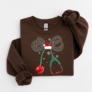 Puede incluir: Una sudadera marr&oacute;n con un dise&ntilde;o de estetoscopio navide&ntilde;o. El estetoscopio tiene un gorro de Pap&aacute; Noel, un lazo con motivos florales y copos de nieve. El dise&ntilde;o est&aacute; impreso en una sudadera de cuello redondo marr&oacute;n.