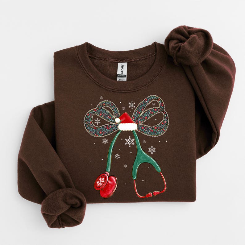 Puede incluir: Una sudadera marr&oacute;n con un dise&ntilde;o de estetoscopio navide&ntilde;o. El estetoscopio tiene un gorro de Pap&aacute; Noel, un lazo con motivos florales y copos de nieve. El dise&ntilde;o est&aacute; impreso en una sudadera de cuello redondo marr&oacute;n.