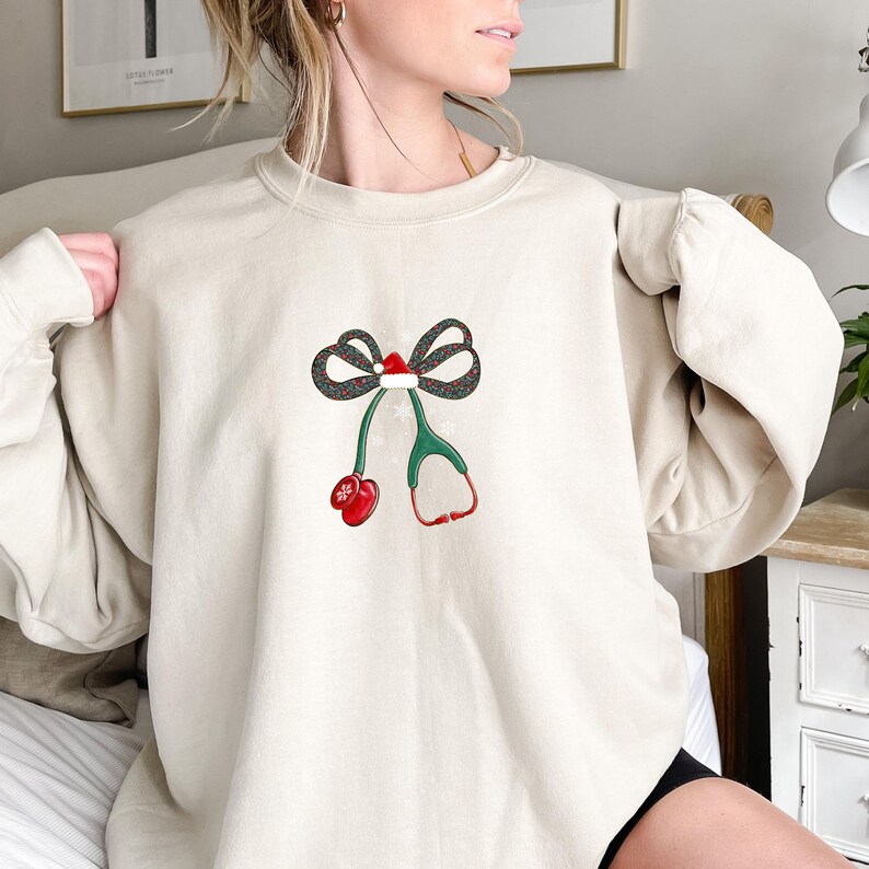 Puede incluir: Sudadera beige con un dise&ntilde;o navide&ntilde;o. El dise&ntilde;o incluye un lazo con un gorro de Pap&aacute; Noel, un estetoscopio verde y un mit&oacute;n rojo. La sudadera est&aacute; hecha de un material suave.