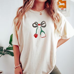 Puede incluir: Camiseta color crema con un dise&ntilde;o festivo. El gr&aacute;fico incluye un lazo con un gorro de Pap&aacute; Noel, copos de nieve, un estetoscopio verde y un mit&oacute;n rojo. El dise&ntilde;o est&aacute; centrado en la parte delantera de la camiseta.