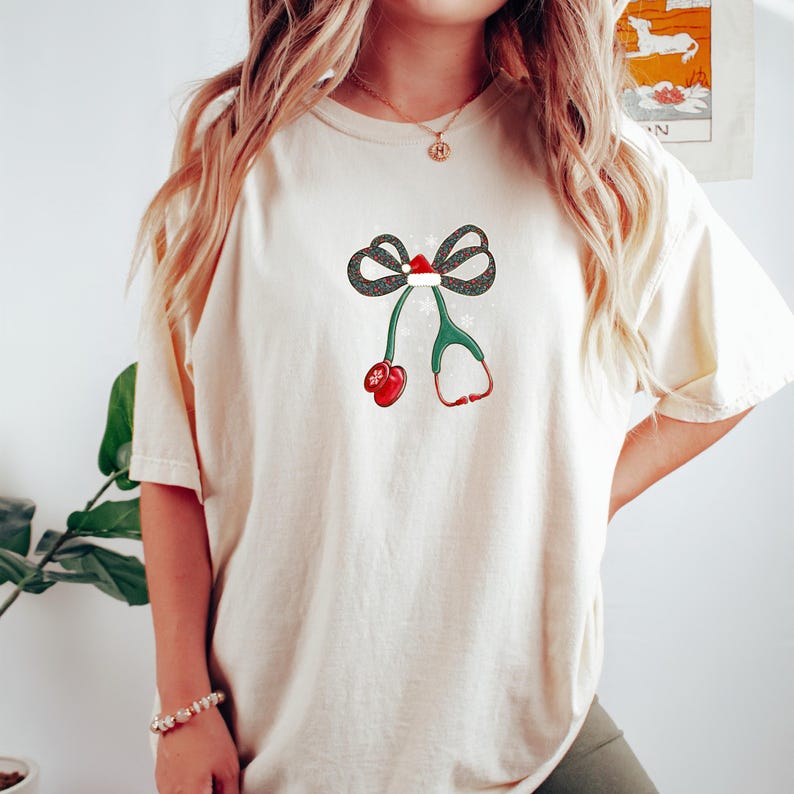 Puede incluir: Camiseta color crema con un dise&ntilde;o festivo. El gr&aacute;fico incluye un lazo con un gorro de Pap&aacute; Noel, copos de nieve, un estetoscopio verde y un mit&oacute;n rojo. El dise&ntilde;o est&aacute; centrado en la parte delantera de la camiseta.
