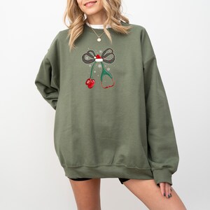 Puede incluir: Una sudadera verde oliva con un dise&ntilde;o navide&ntilde;o. El gr&aacute;fico incluye un lazo con un gorro de Pap&aacute; Noel, copos de nieve, un estetoscopio y mitones rojos. El dise&ntilde;o est&aacute; centrado en la parte delantera de la sudadera.