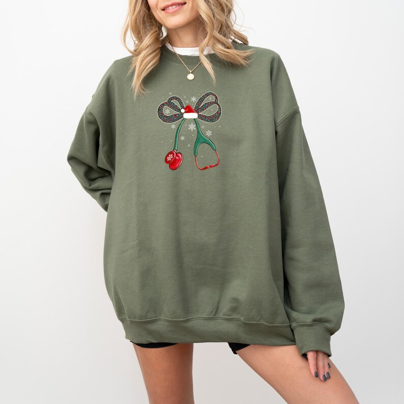 Puede incluir: Una sudadera verde oliva con un dise&ntilde;o navide&ntilde;o. El gr&aacute;fico incluye un lazo con un gorro de Pap&aacute; Noel, copos de nieve, un estetoscopio y mitones rojos. El dise&ntilde;o est&aacute; centrado en la parte delantera de la sudadera.