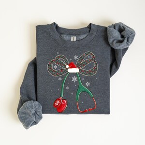 Puede incluir: Sudadera gris oscuro con un dise&ntilde;o navide&ntilde;o. El gr&aacute;fico presenta un lazo con un gorro de Pap&aacute; Noel, copos de nieve y un estetoscopio. El estetoscopio es verde con detalles rojos, creando un tema m&eacute;dico festivo.
