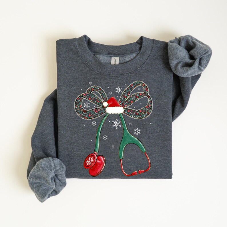 Puede incluir: Sudadera gris oscuro con un dise&ntilde;o navide&ntilde;o. El gr&aacute;fico presenta un lazo con un gorro de Pap&aacute; Noel, copos de nieve y un estetoscopio. El estetoscopio es verde con detalles rojos, creando un tema m&eacute;dico festivo.