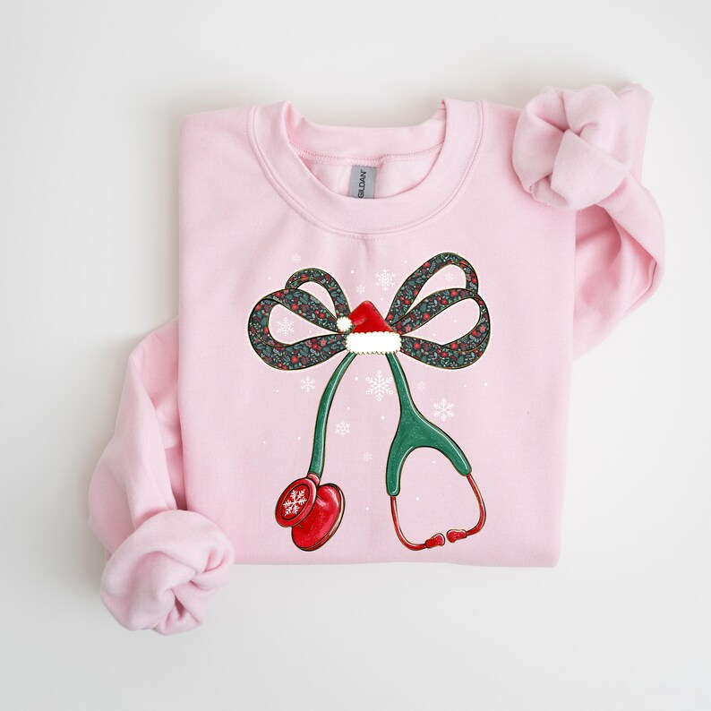 Puede incluir: Una sudadera rosa claro con un dise&ntilde;o de estetoscopio navide&ntilde;o. El estetoscopio es verde con detalles rojos, incluyendo un gorro de Pap&aacute; Noel en el lazo y detalles de copos de nieve. El lazo tiene un estampado floral.