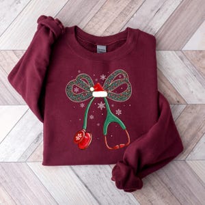 Puede incluir: Sudadera burdeos con un dise&ntilde;o de lazo navide&ntilde;o, un gorro de Pap&aacute; Noel y un estetoscopio. El lazo es verde con detalles rojos y blancos, y hay copos de nieve dispersos. El estetoscopio es verde y rojo.