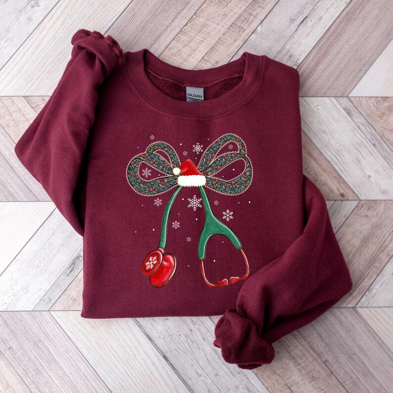 Puede incluir: Sudadera burdeos con un dise&ntilde;o de lazo navide&ntilde;o, un gorro de Pap&aacute; Noel y un estetoscopio. El lazo es verde con detalles rojos y blancos, y hay copos de nieve dispersos. El estetoscopio es verde y rojo.