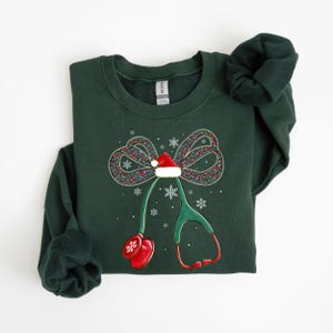 Puede incluir: Sudadera verde oscuro con un dise&ntilde;o navide&ntilde;o. El dise&ntilde;o incluye un lazo con un gorro de Pap&aacute; Noel, copos de nieve y un estetoscopio. El estetoscopio tiene detalles en rojo y un mit&oacute;n rojo. La sudadera est&aacute; doblada.
