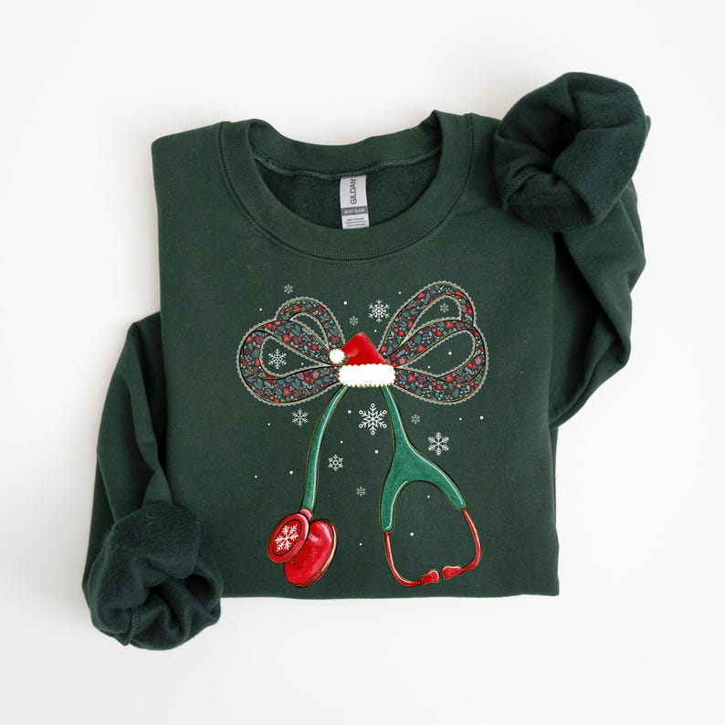 Puede incluir: Sudadera verde oscuro con un dise&ntilde;o navide&ntilde;o. El dise&ntilde;o incluye un lazo con un gorro de Pap&aacute; Noel, copos de nieve y un estetoscopio. El estetoscopio tiene detalles en rojo y un mit&oacute;n rojo. La sudadera est&aacute; doblada.