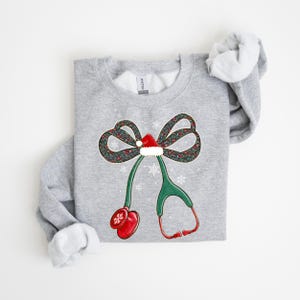 Puede incluir: Sudadera gris claro con un dise&ntilde;o festivo. El dise&ntilde;o incluye un lazo con un estampado floral, un gorro de Pap&aacute; Noel y un estetoscopio. El estetoscopio tiene auriculares rojos y una pieza tor&aacute;cica verde. Los copos de nieve est&aacute;n dispersos alrededor del dise&ntilde;o.