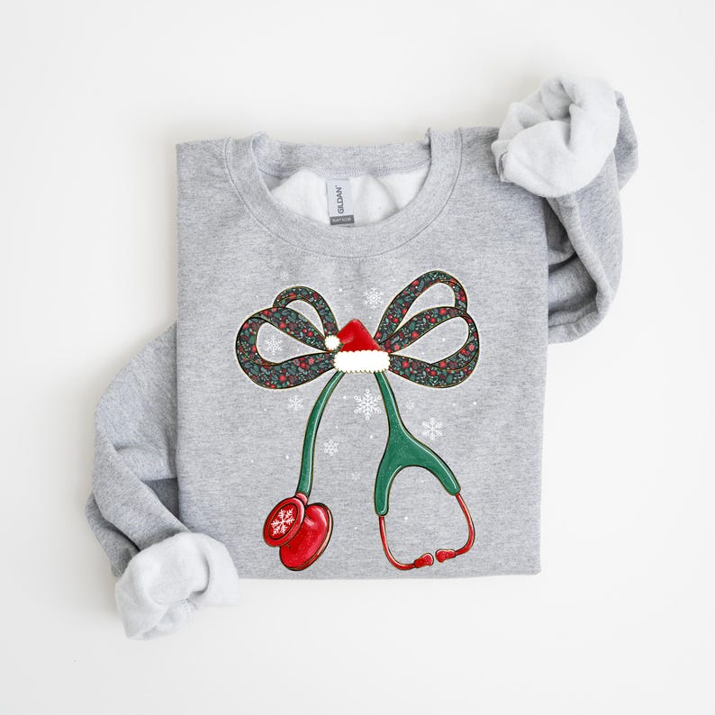Puede incluir: Sudadera gris claro con un dise&ntilde;o festivo. El dise&ntilde;o incluye un lazo con un estampado floral, un gorro de Pap&aacute; Noel y un estetoscopio. El estetoscopio tiene auriculares rojos y una pieza tor&aacute;cica verde. Los copos de nieve est&aacute;n dispersos alrededor del dise&ntilde;o.