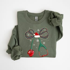 Puede incluir: Una sudadera verde oliva con un dise&ntilde;o navide&ntilde;o. El dise&ntilde;o incluye un lazo con un gorro de Pap&aacute; Noel, copos de nieve y un estetoscopio. El lazo tiene un estampado floral y el estetoscopio es verde y rojo.