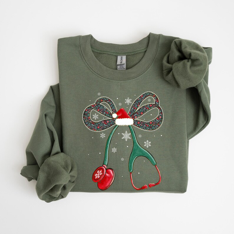 Puede incluir: Una sudadera verde oliva con un dise&ntilde;o navide&ntilde;o. El dise&ntilde;o incluye un lazo con un gorro de Pap&aacute; Noel, copos de nieve y un estetoscopio. El lazo tiene un estampado floral y el estetoscopio es verde y rojo.