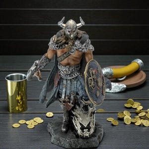 May include: A detailed figurine of a Viking warrior, in ornate armour and a horned helmet, holding a shield. The figure stands on a stone base with a skull, accompanied by a golden cup, coins, and a drinking horn. The overall theme is Norse mythology.