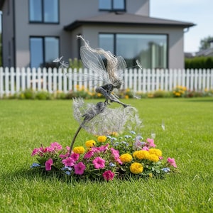 May include: A whimsical metal fairy sculpture with wire wings and flowing hair, perched atop a dandelion. The fairy is surrounded by a vibrant bed of pink, yellow, and blue flowers, set against a green lawn and a white picket fence.