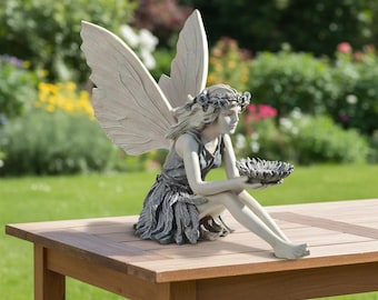 Könnte beinhalten: Eine hellgraue Feenstatue mit großen, texturierten Flügeln, die einen Blumenkranz und ein Kleid mit Blattmuster trägt. Die Figur sitzt und hält eine kleine, flache, blütenförmige Schale in beiden Händen.