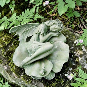 以下が含まれることがあります： 大きな葉の上に横たわる、穏やかな緑色の妖精の像。小さなカエルがそばにいます。妖精は、なびく髪と繊細な翼で描かれており、庭の装飾品として魅力的です。像は緑に囲まれています。