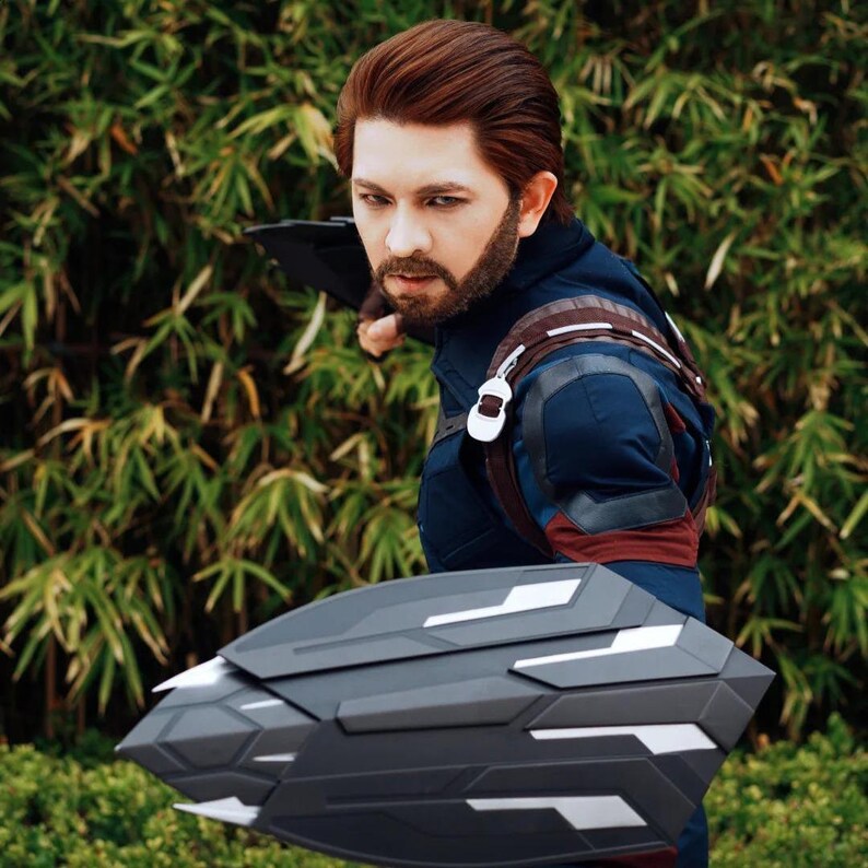 May include: A person in a Captain America costume, holding a gray and black shield with white accents. The costume is primarily navy blue with red and brown details. The background is a lush green foliage.