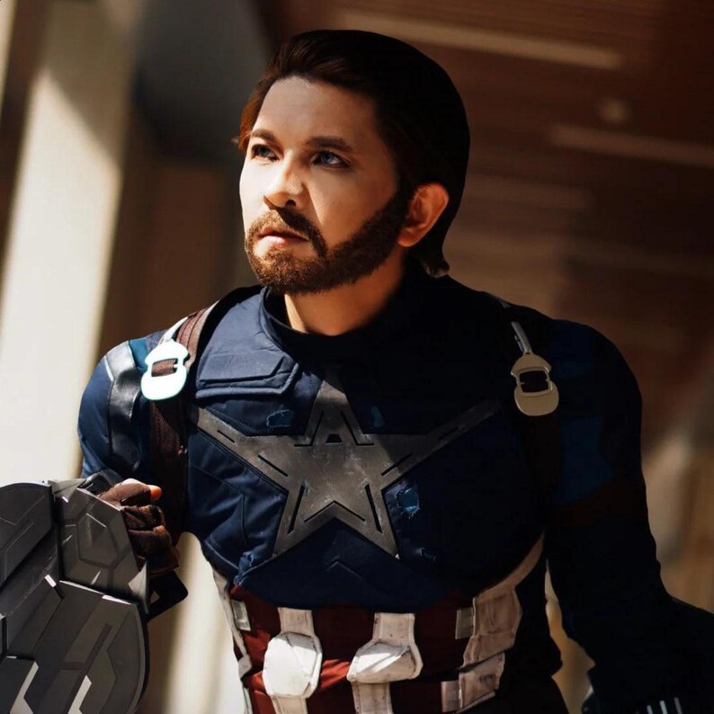 May include: A person dressed as Captain America, wearing a blue and red costume with a white star on the chest. The costume includes a shield and a brown leather strap. The person has brown hair and a beard.