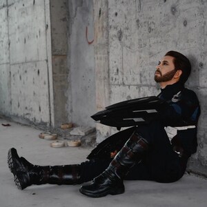 May include: A person in a Captain America costume, seated against a concrete wall. The costume includes a navy blue and white suit, black boots, and a black shield. The person has a brown beard and is looking upwards.