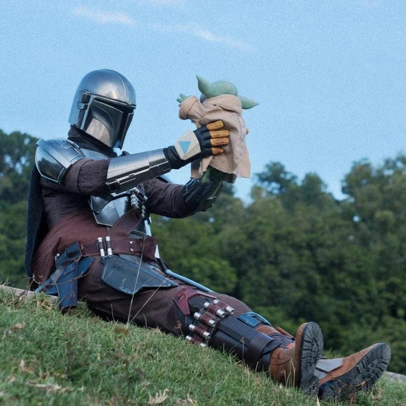 May include: A person in a Mandalorian costume, complete with a silver helmet and armor, holding a Grogu doll. The person is seated on a grassy hill with a forest in the background. The costume features brown and black elements.
