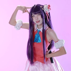 Peut inclure: Un costume de cosplay avec un haut rouge, une cravate bleue et une jupe rose. Le personnage a de longs cheveux violets, une coiffe décorative et des manches bouffantes blanches. Le fond est de couleur rose uni.