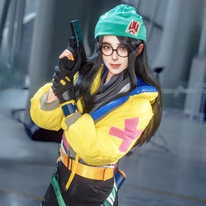 May include: A person in a cosplay outfit, wearing a yellow jacket with blue and pink accents, a green beanie, and round glasses. They are holding a black toy gun. The outfit includes black gloves and a utility belt.