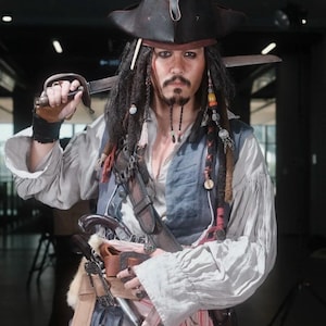 Könnte beinhalten: Eine Person, die als Captain Jack Sparrow verkleidet ist und ein Schwert hält. Sie trägt einen schwarzen Dreispitz, ein weißes Hemd, eine blaue Weste und hat Dreadlocks mit Perlen. Ein Ledergürtel mit Scheide und Pistolen ist sichtbar.