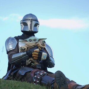 May include: A person in Mandalorian armor holding a Grogu doll. The armor is silver and black, with a helmet and shoulder pads. Grogu is a small, green alien with large ears, wrapped in a tan blanket. The background is a blue sky.
