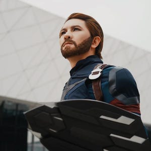 May include: A person in a Captain America costume, looking up with a determined expression. The costume is primarily navy blue with red and white accents, and a brown leather strap. The shield is black with silver details.