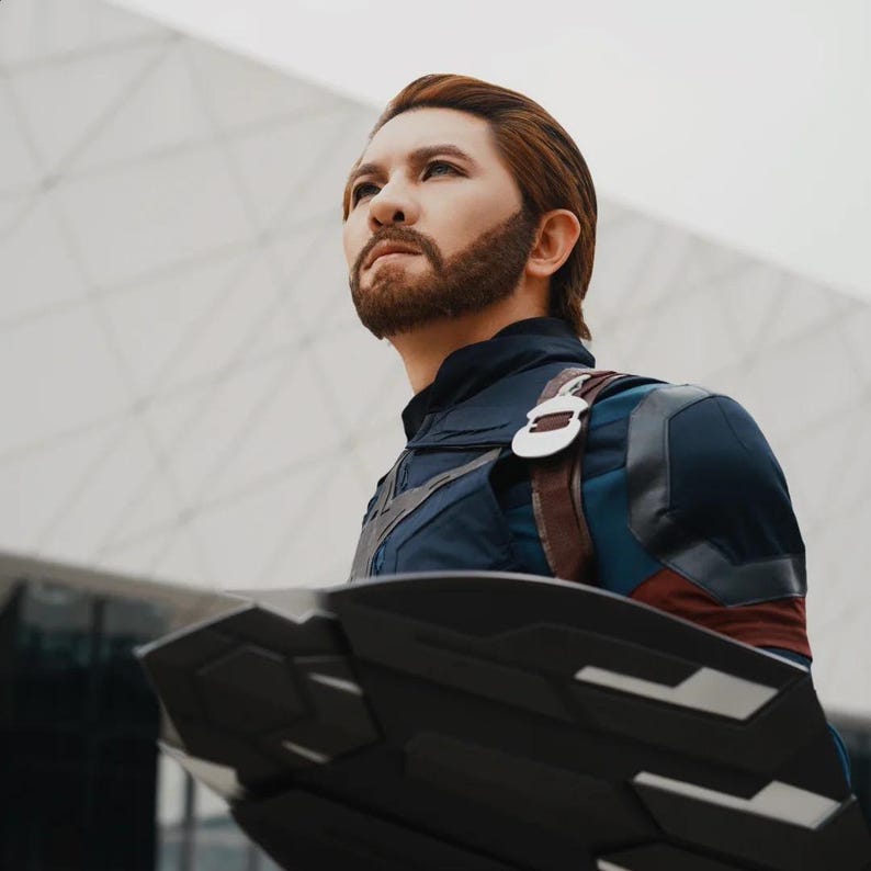 May include: A person in a Captain America costume, looking up with a determined expression. The costume is primarily navy blue with red and white accents, and a brown leather strap. The shield is black with silver details.