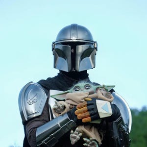May include: A person in a silver Mandalorian helmet and armor holds a Grogu doll. The doll is wrapped in a tan blanket. The background is a clear, light blue sky. The Mandalorian's armor has a dark grey cape.