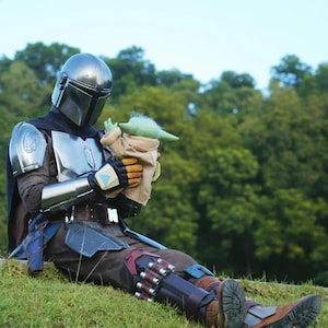 May include: A person in a Mandalorian costume sits on a grassy hill, holding a Grogu doll. The costume features a silver helmet, chest plate, and detailed armor. The Grogu doll is green with large ears and is wrapped in a tan blanket.