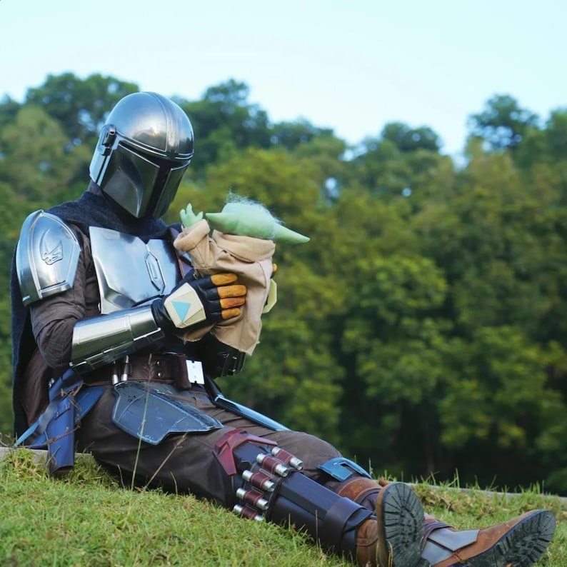 May include: A person in a Mandalorian costume sits on a grassy hill, holding a Grogu doll. The costume features a silver helmet, chest plate, and detailed armor. The Grogu doll is green with large ears and is wrapped in a tan blanket.